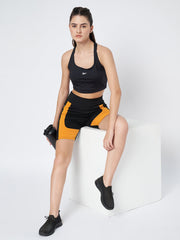 Waylene's sports model posing on a white block in high-waist black and orange sports shorts, paired with a black sports bra and holding a water bottle, perfect for running & yoga.