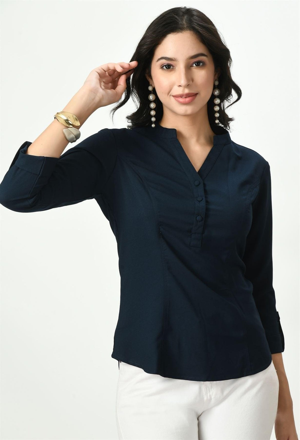 A woman with her hand on her hip, wearing a dark navy blue Waylene button-up top, paired with white pants, against a plain background.